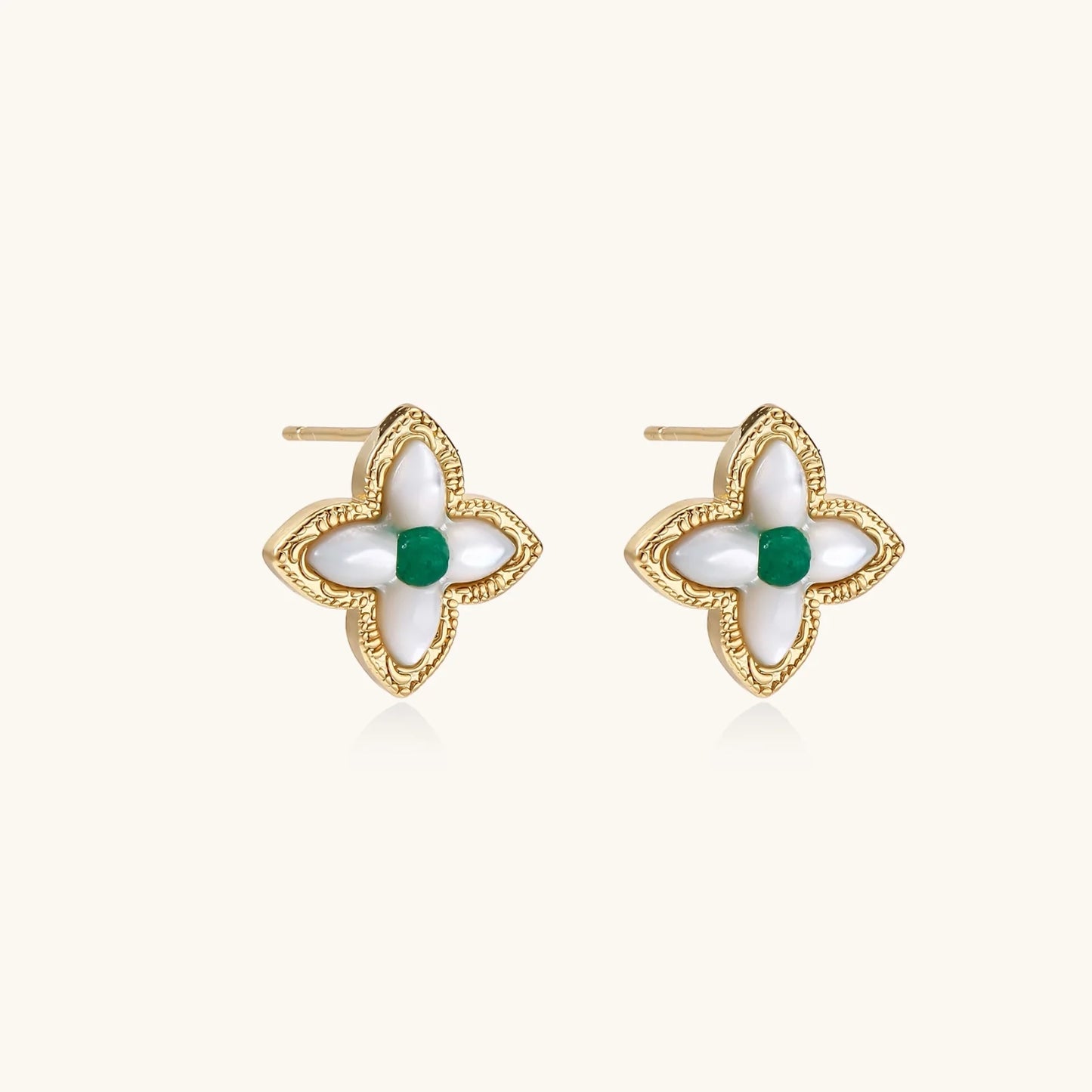 Gold quatrefoil stud earrings with white mother-of-pearl petals and emerald green gemstone center on beige background.