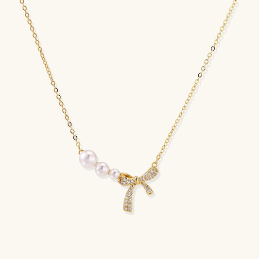 Delicate gold necklace with three white pearls and crystal-embellished bow pendant on cream background