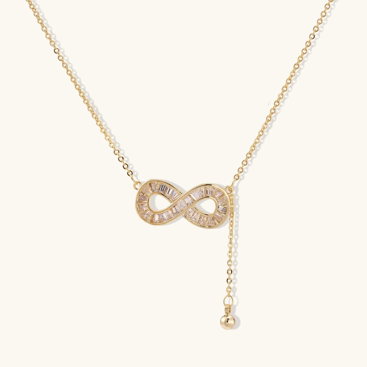 Gold infinity pendant with baguette crystals on adjustable chain against cream background.